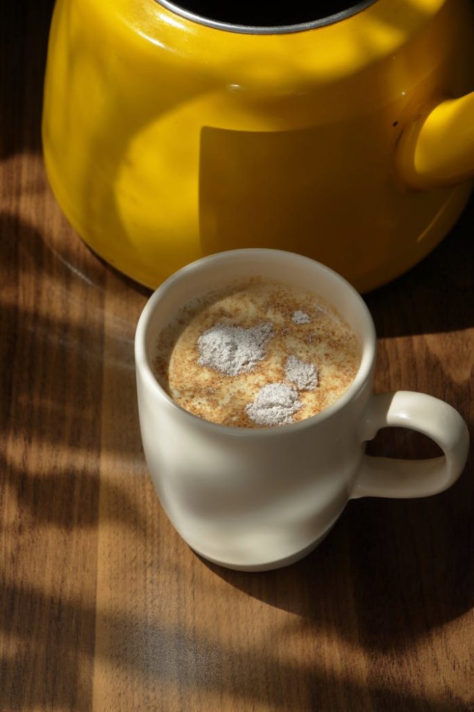 A warm beverage in a ceramic mug under sunlight with a yellow teapot.